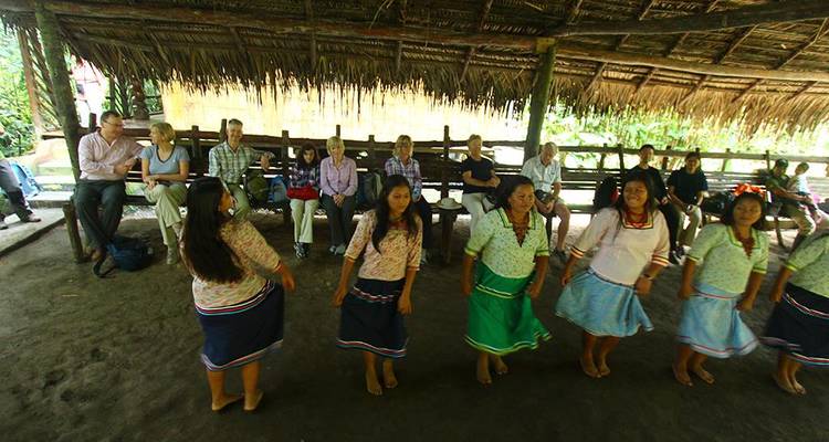 Indigenous women perform a traditional dance for seated tourists under a thatched community shelter.