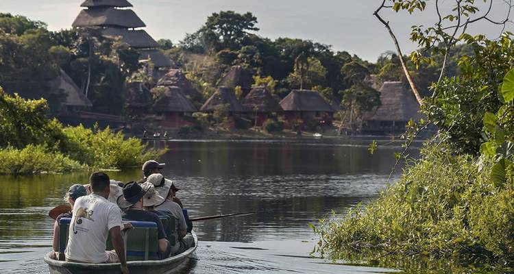 Tourists ride in a narrow canoe across a reflective rainforest lake toward thatched lodge huts.