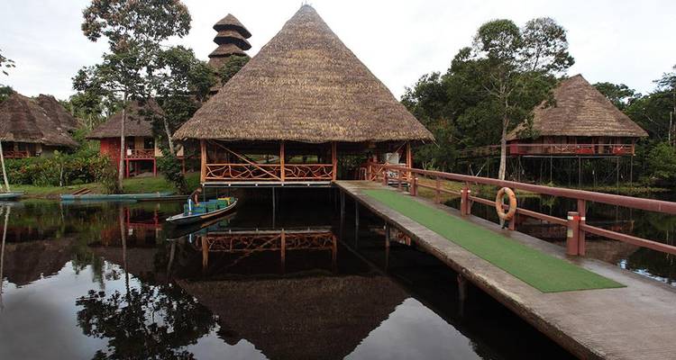 Thatched-roof lodge complex reflected perfectly in dark still water, linked by a wooden bridge.