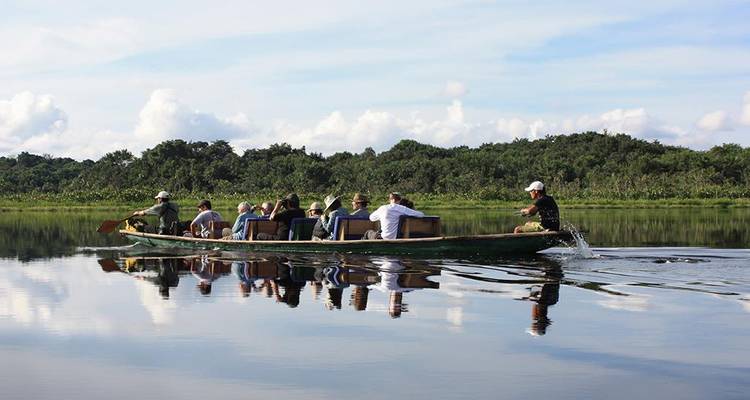 A motorized wooden canoe carries tourists across a mirror-like river with lush banks reflecting on the water.