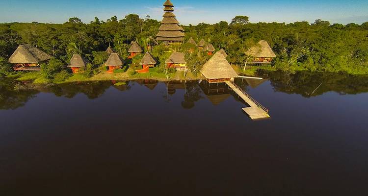 Drone view of a lakeside eco-lodge with multiple thatched huts and a tall central tower amid dense jungle.