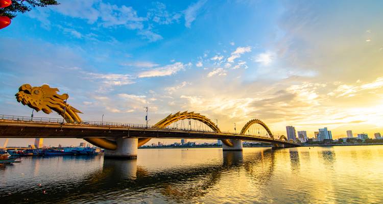 Een drakenbrug over een rivier bij zonsondergang met een stadsgezicht.