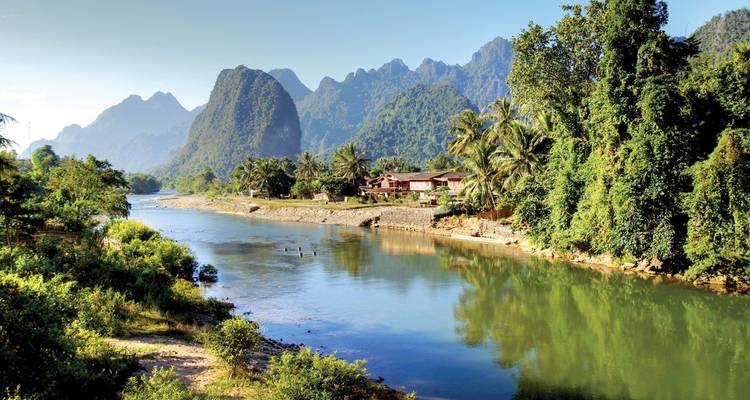 Un fleuve tranquille serpente devant des maisons tropicales et des montagnes de calcaire spectaculaires sous un ciel bleu dégagé.