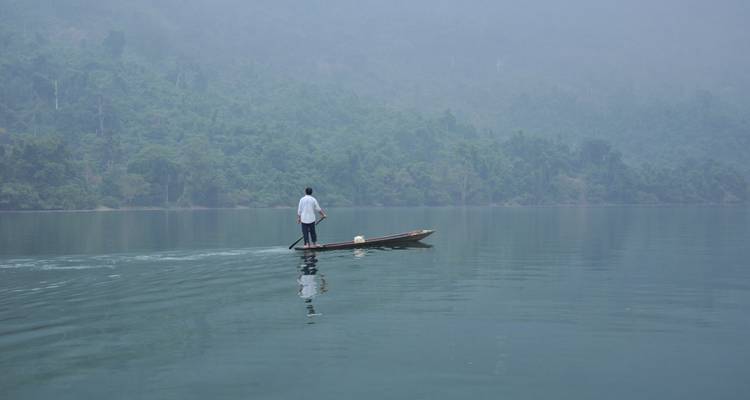 Hombre solitario de pie en una canoa de madera estrecha remando a través de un lago brumoso