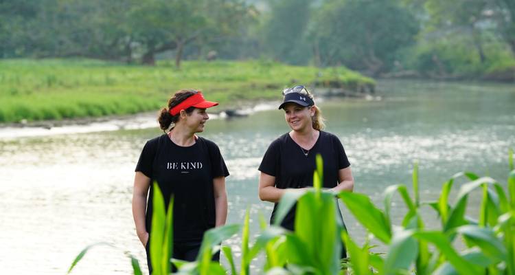 Two women chat while walking beside a quiet river and green riverside crops.