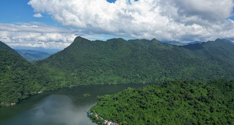 Wide aerial of forested mountains surrounding a calm lake with scattered floating houses.