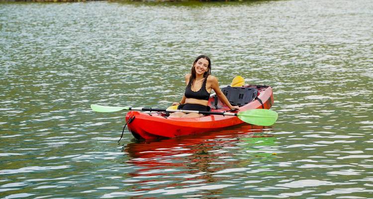 Female traveller paddles a red kayak on shimmering lake water while smiling at the camera.