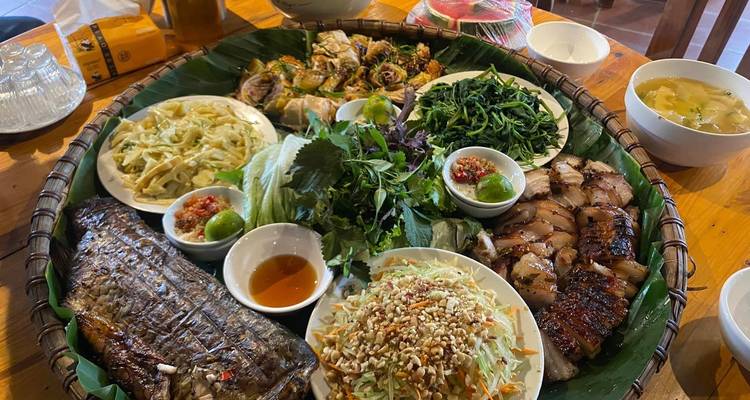 Large bamboo tray filled with grilled fish, spring rolls, greens and other local specialties on a wooden table.