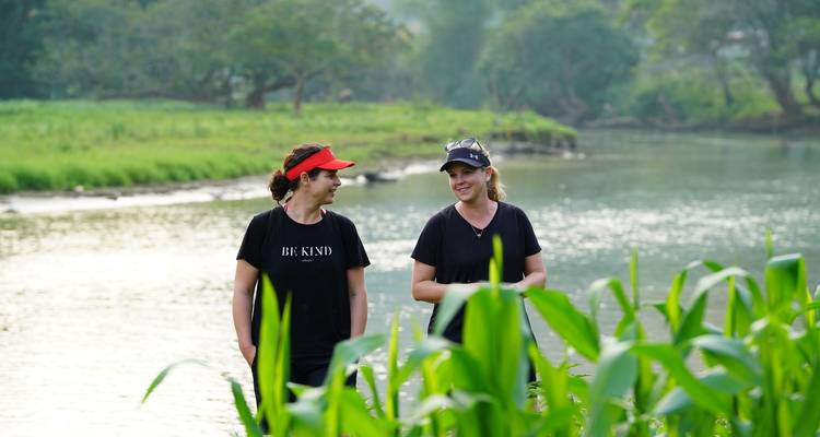 Two women walk beside a tranquil river bordered by tall green corn plants.
