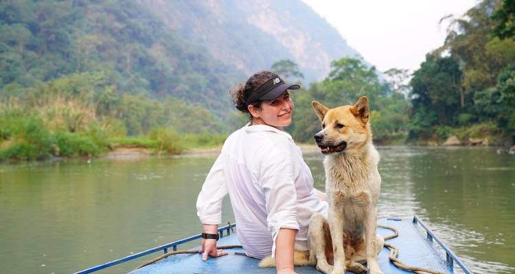 Traveller sits with a friendly dog on the bow of a blue boat cruising a misty river gorge.