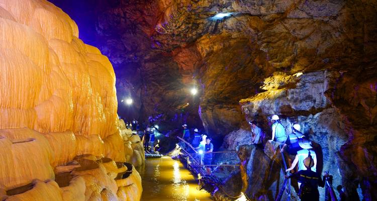 Illuminated limestone cave with amber and blue lighting where visitors walk on a raised boardwalk.