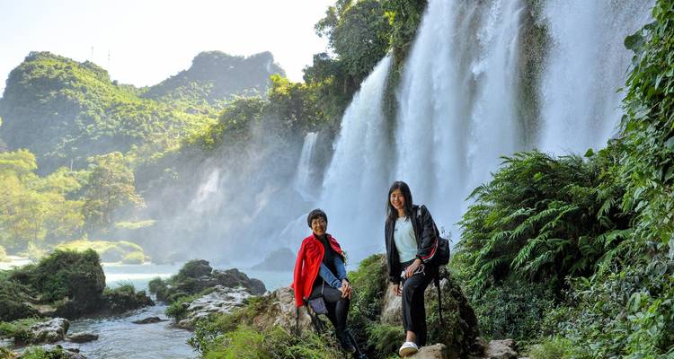 Two female travellers posing on rocks beside a tall, misty waterfall surrounded by lush green cliffs.