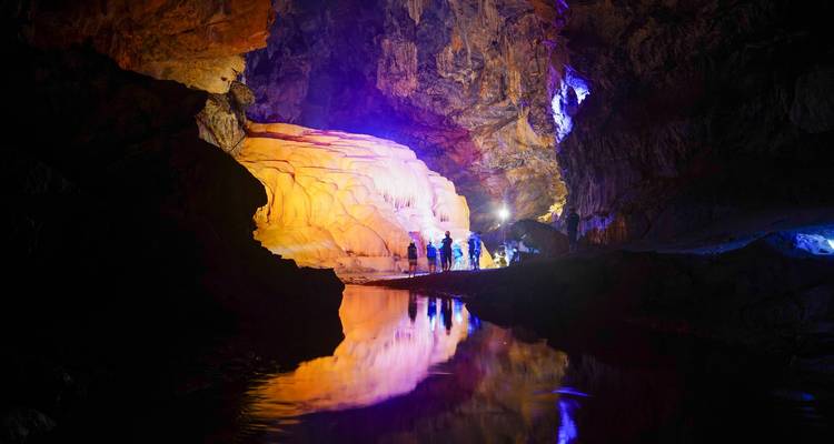 Illuminated cave interior with colourful lights reflecting on an underground stream while a small group explores.