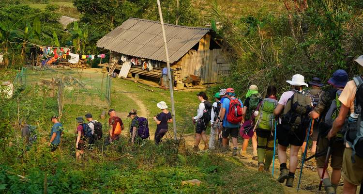 Hiking group with backpacks follows a dirt trail past a wooden rural house and tropical greenery.