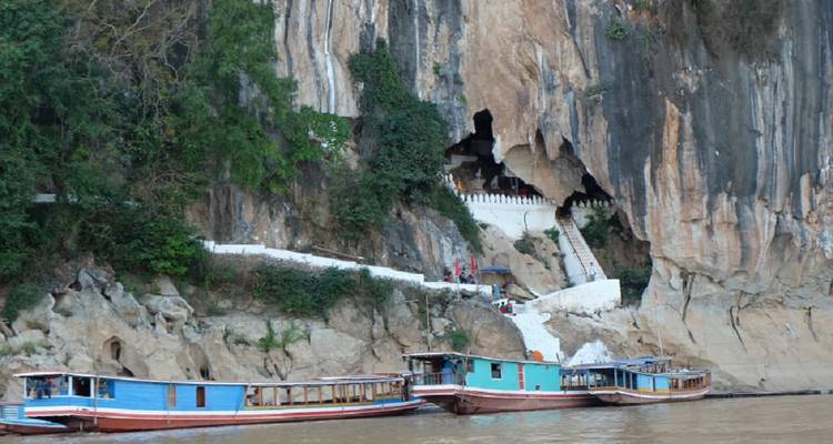Long-tail boats moored at the mouth of Pak Ou Caves carved into a limestone cliff above the Mekong River.