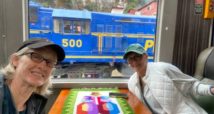 Deux femmes souriantes à l'intérieur d'un wagon de train avec une locomotive bleue PeruRail à l'extérieur de la fenêtre