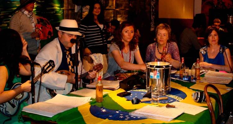 Des musiciens et des convives sont assis autour de tables recouvertes d'un drapeau brésilien dans un pub ou café animé.
