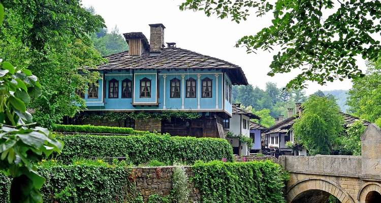Maison balkanique traditionnelle bleue avec balcons en bois entourée d'une végétation luxuriante et d'un pont en pierre.