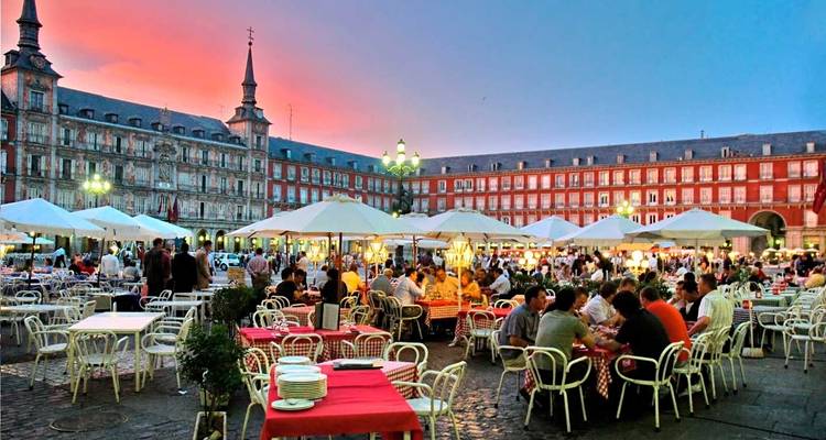 Scène de dîner animée en soirée sur la Plaza Mayor de Madrid avec parasols, tables et foules joyeuses.