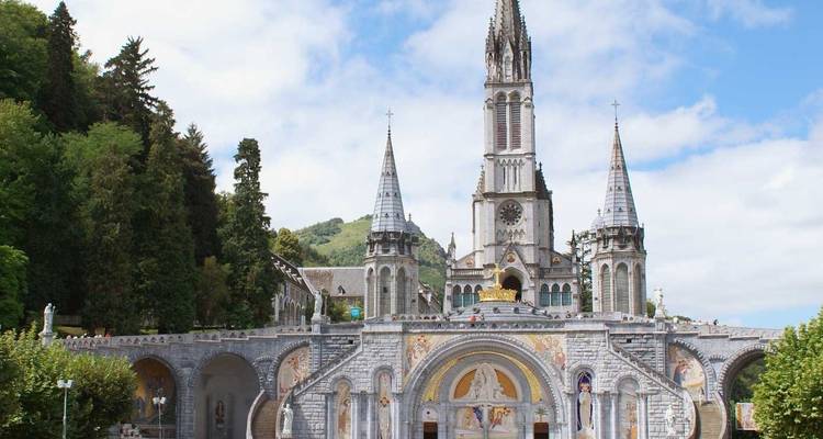La Basilique ornée de Notre-Dame de Lourdes avec sa façade en mosaïque et ses deux flèches dressées contre les collines boisées.