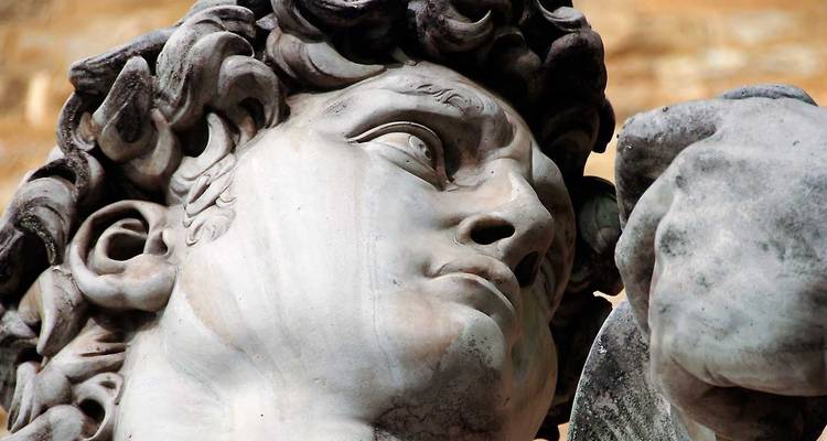 Vista de cerca del rostro de mármol y cabello rizado de la estatua del David de Miguel Ángel contra un fondo de piedra cálida.