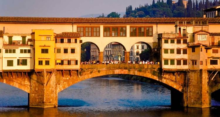 El Ponte Vecchio iluminado en dorado que se extiende sobre el río Arno con multitudes de visitantes de pie a lo largo de su centro al anochecer.