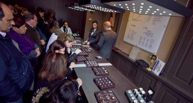 Grupo de turistas reunidos dentro de una joyería examinando bandejas de relojes y anillos.