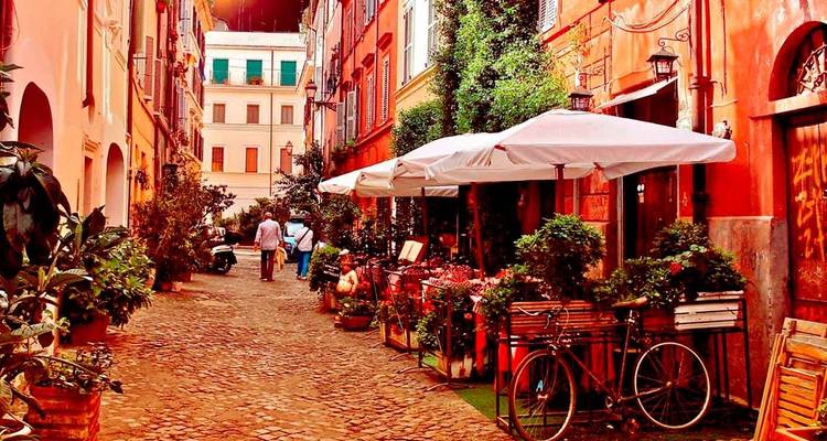Callejón romano angosto y colorido lleno de cafes, bicicletas y plantas bajo la cálida luz de la tarde.