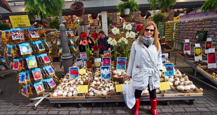 Mujer sonriente posa en el mercado de flores de Ámsterdam rodeada de bulbos de tulipán y exhibiciones coloridas.