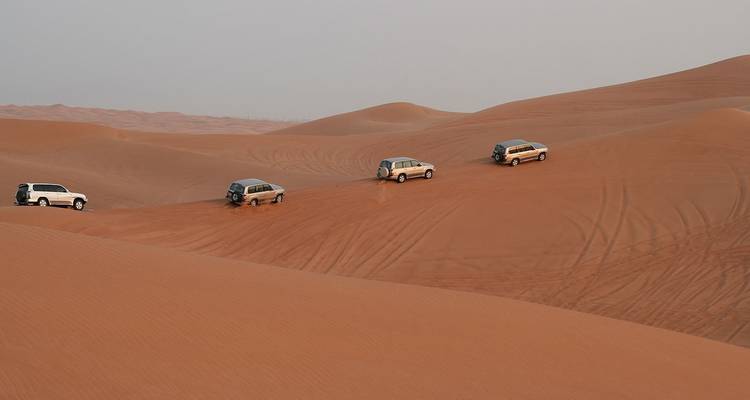 Vier Geländewagen fahren im Gänsemarsch über weite, gewellte Wüstendünen und hinterlassen geschwungene Spuren im Sand.