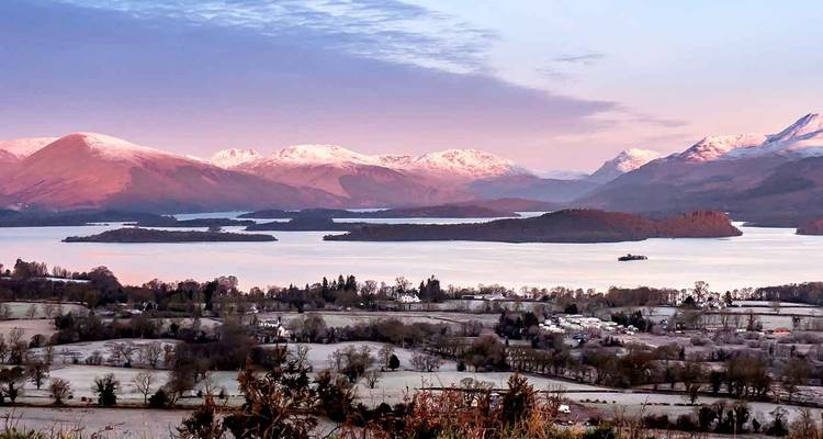 Les montagnes aux sommets enneigés brillent en rose au-dessus d'un vaste lac parsemé d'îlots boisés et d'un paysage rural glacé au premier plan.