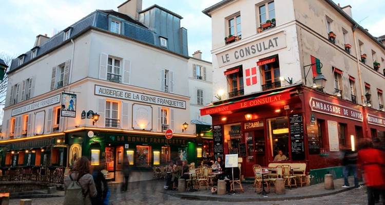 Coin animé de Montmartre avec les cafés Le Consulat et La Bonne Franquette illuminés au crépuscule.
