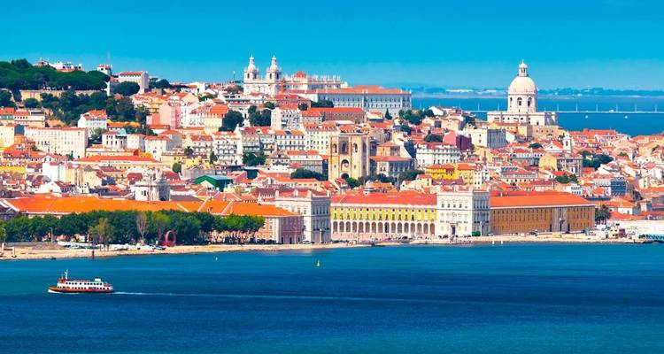 Colourful hillside cityscape of Lisbon stretching along the Tagus waterfront under a bright blue sky.