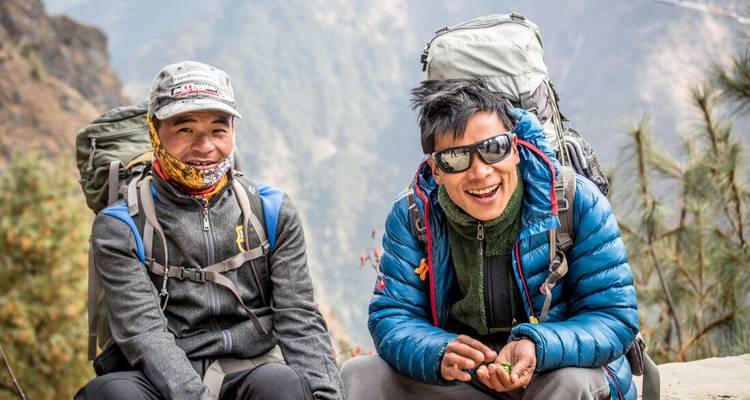 Dos guías nepalíes con mochilas grandes sonríen mientras descansan en un sendero de montaña.