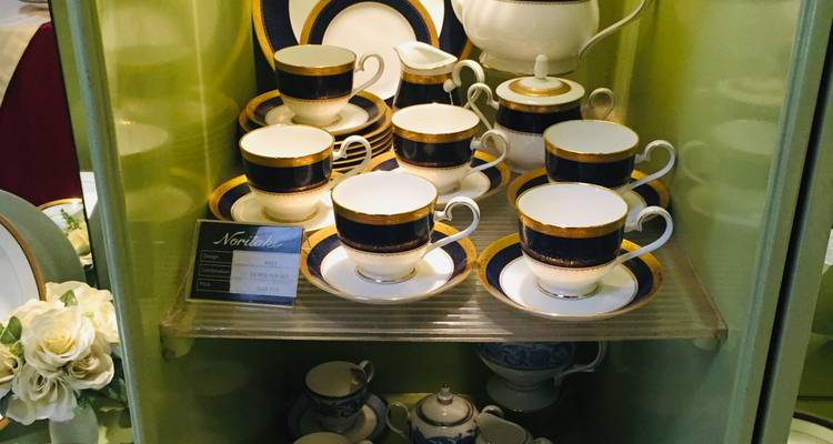 Vitrine d'exposition présentant de fines tasses à thé Noritake avec des bordures bleues et dorées dans une salle d'exposition d'usine.