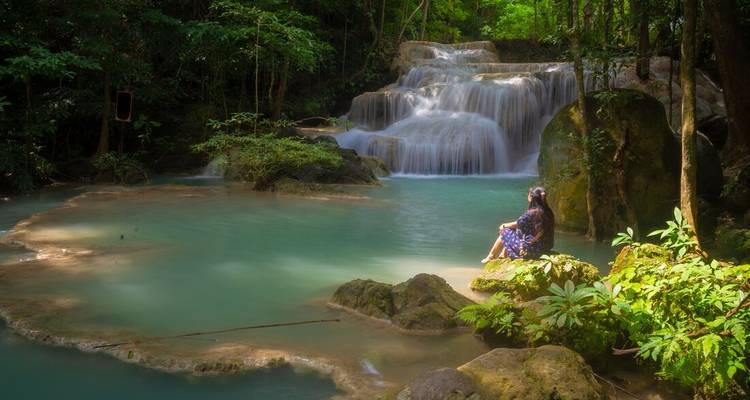 Wasserfall, der in einen ruhigen Teich in einem Wald fließt, mit einer Person, die am Wasser sitzt.