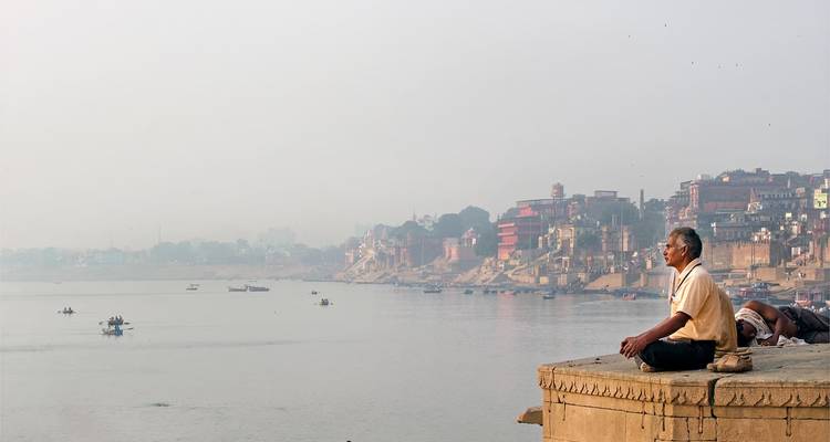 An elderly man meditating on the ghats of Varanasi, with riverboats in the background.