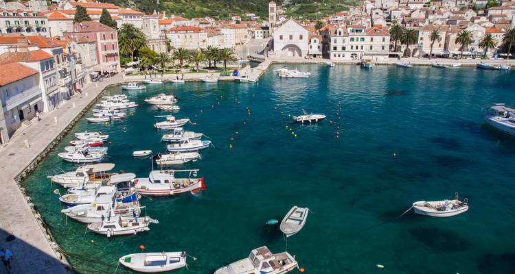 Scène portuaire de la ville de Hvar avec de petits bateaux amarrés sur une eau turquoise cristalline à côté de bâtiments aux couleurs pastel et une église sur la colline.