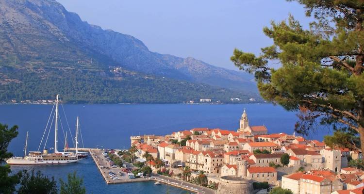 Vue panoramique de la vieille ville de Korčula avec ses toits en terre cuite, le clocher de l'église et les yachts ancrés face aux montagnes imposantes et aux eaux bleu profond.