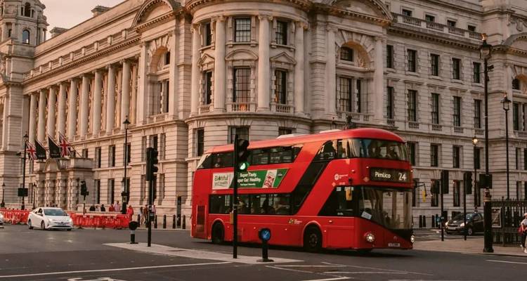 Un autobus à impériale rouge roulant devant des bâtiments en pierre blanche ornée sur une avenue londonienne à la lumière dorée du soir.