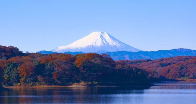 Le mont Fuji couronné de neige s'élève derrière une forêt d'automne colorée se reflétant dans un lac tranquille.