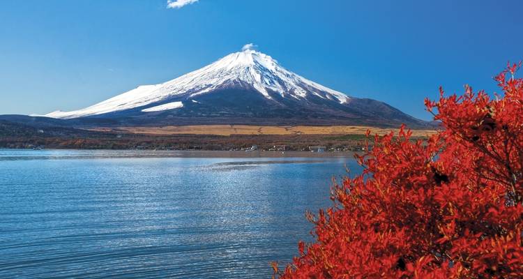 De met sneeuw bedekte berg Fuji die oprijst boven een kalm blauw meer omzoomd door levendig rood herfstgebladerte.