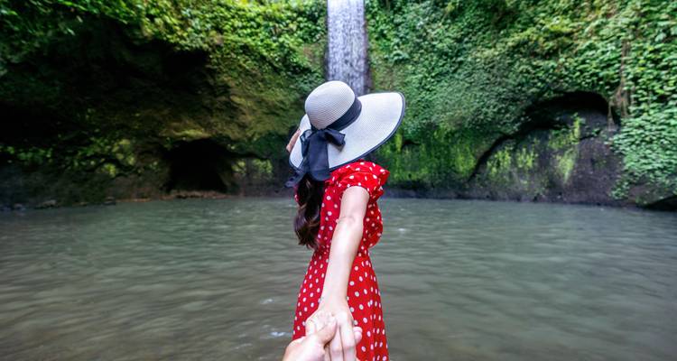 Person in a red dress holding someone's hand in front of a waterfall.
