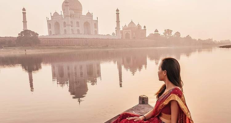 Een vrouw in een traditionele sari zit in een klein bootje en kijkt over een rustige rivier naar het Taj Mahal dat in het zachte ochtendlicht weerspiegelt.