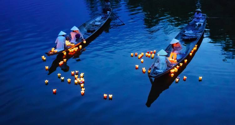 Nachtelijke rivier lantaarnceremonie met deelnemers met kegelvormige hoeden die gloeiende kaarsen plaatsen vanuit twee houten boten.