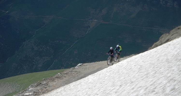 Deux vététistes gravissent une pente neigeuse abrupte avec des pics verts accidentés au loin.