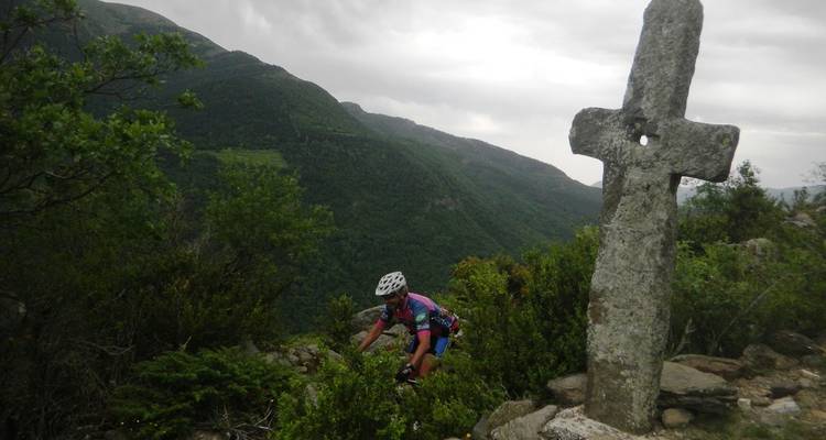Un cycliste négocie un sentier rocheux près d'une croix de pierre ancienne surplombant une vallée escarpée.
