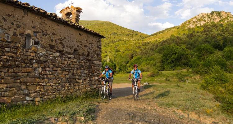 Deux vététistes roulent sur un chemin de terre à côté d'une ancienne chapelle en pierre située dans des collines verdoyantes vallonnées.