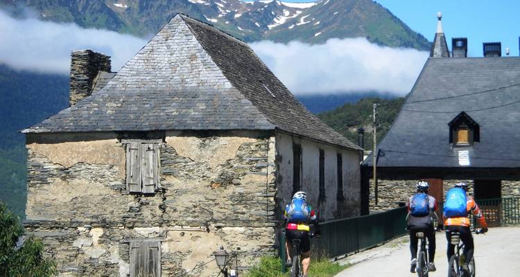 Trois vététistes passent devant des cottages de pierre rustiques dans un hameau de haute montagne.