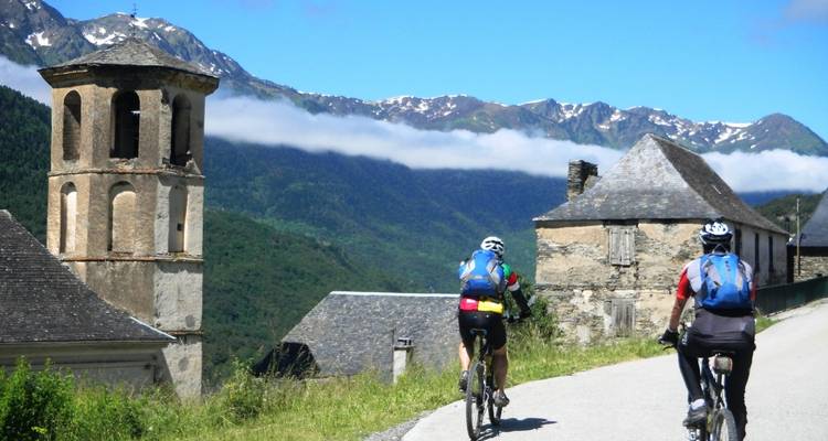 Deux cyclistes pédalent devant un clocher de pierre rustique avec les montagnes pyrénéennes aux sommets enneigés en arrière-plan.
